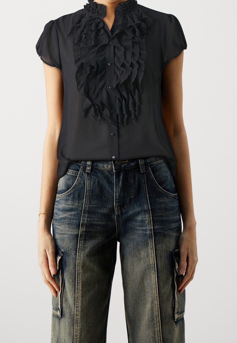 Personne portant un chemisier noir à manches courtes avec des volants et un jean cargo en denim bleu délavé taille haute avec poches avant et latérales.
