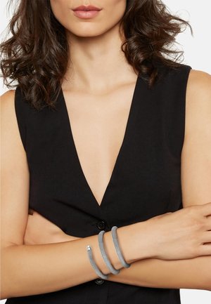 Femme aux cheveux bruns bouclés porte un haut noir sans manches à col en V et un bracelet en spirale argentée texturée autour de ses avant-bras croisés.