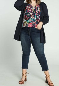 Cárdigan negro sobre una blusa de colores y estampados con escote en V; combinado con unos vaqueros oscuros de longitud hasta el tobillo y sandalias con tacones de tiras marrones.