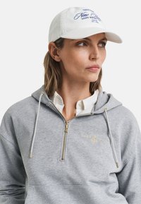 Hallhõlmik hall kapuutsiga, millel on kuldne tikand ja reguleeritav kapuuts. Valge müts sinise tikandiga. Mudel kannab kaelusega särke.