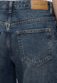 Κοντινή λήψη της πίσω τσέπης και της μέσης μπλε τζιν με καφέ δερμάτινη ετικέτα με την επιγραφή "SAMSØE SAMSØE."