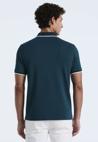 Dunkelgrünes Polohemd aus texturiertem Stoff, mit kurzen Ärmeln und weißen Akzenten am Kragen und den Ärmelsäumen. Rückansicht gezeigt.