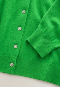 Cardigan verde, cu textură netedă, șapte nasturi argintii, manșete și tiv cu trotuar. Material moale, ușor, cu un design de culoare solidă.