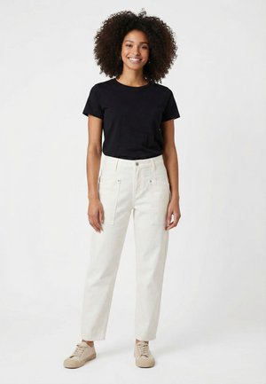 Lachende vrouw met krullend haar, draagend een zwart T-shirt, witte hoog getailleerde broek met voorzakken en beige sneakers, staand op een witte achtergrond.