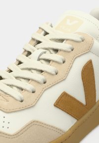 Sneaker bianca e beige con tomaia in pelle testurizzata, design con lacci e dettaglio del logo a "V" in suede marrone sul lato. Suola in gomma.