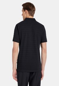 Mężczyzna stojący tyłem, ubrany w czarną koszulkę polo z krótkim rękawem i czarne spodnie, na jednolitym jasnym tle.