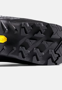 Zwarte rubberen schoenzool met diepe geometrische profielen en een geel Vibram-logo vlak bij de hiel.