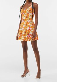 Nainen, joka pukeutuu olkaimettomaan, muotoon sovitettuun kesämekkoon, jossa on oranssi, keltainen ja valkoinen kukkakuosi, seisoo valkoista taustaa vasten. 