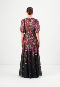 Vrouw met een bun in haar haar, gekleed in een zwarte maxi-jurk met kleurrijke bloemmotieven en pof korte mouwen, staand met haar rug naar de kijker.