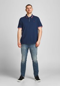 Marineblauwe polo met een rood-wit gestreepte kraag, korte mouwen en drie knopen bij de hals, gecombineerd met lichtblauwe jeans en zwarte sneakers.