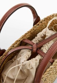 Tkana słomiana torba z brązowymi skórzanymi uchwytami i dodatkami, wyposażona w beżową, materiałową podszewkę ze ściągaczem zabezpieczoną u góry.