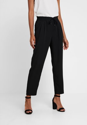 Vrouw draagt zwarte, hooggetailleerde taps toelopende broek met trekkoord en zwarte sandaal met blokhak en enkelriem.