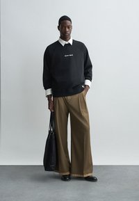 Černá mikina se bílým logem, přelívaná přes bílou košilovou košili. Béžové široké kalhoty a černé boty. Černá kabelka v ruce.