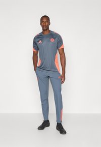 Grå atletisk trøje med orange accenter og logo, parret med matchende grå bukser med tre orange striber og en slim fit design.