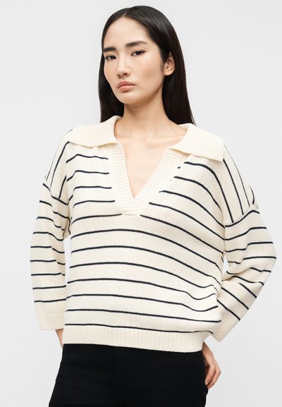 Γυναίκα με μακριά σκουρόχρωμα μαλλιά, φοράει λευκό πλεκτό πουλόβερ με μαύρες οριζόντιες ρίγες και βαθύ V λαιμόκοψη, στέκεται με το ένα χέρι στη μέση.