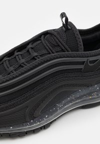Chaussure de sport noire avec une tige en textile et synthétique superposée, semelle épaisse translucide mouchetée et tirette sur la languette.