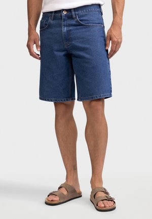 Man die blauwe spijkershorts en bruine suède sandalen draagt, staand tegen een effen achtergrond.