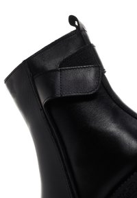 Bota de cuero negro con una textura suave, que presenta una correa de Velcro, un diseño de tobillo curvado y un detalle de costura en contraste.