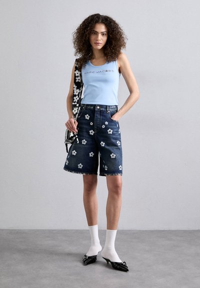 Shorts de denim con bordado floral en blanco, una camiseta sin mangas azul con logo y accesorios negros. La modelo lleva calcetines blancos y zapatos de punta.