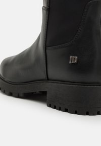 Bota de cuero negra con un acabado texturizado, que presenta una suela de goma resistente y un detalle metálico en el costado, destacando un diseño elegante.