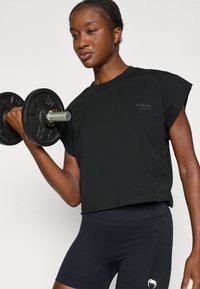 Schwarzes, kurzes T-Shirt mit kurzen Ärmeln und dezentem Branding, kombiniert mit enganliegenden schwarzen Shorts mit strukturierten Akzenten, während ein Kurzhantel gehalten wird.