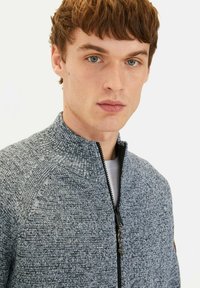 Grau melierte, strukturierte Zip-Up-Jacke mit hohem Kragen, ausgestattet mit einem dezenten Muster und einem schwarzen Reißverschluss. Getragen über einem hellen Hemd.
