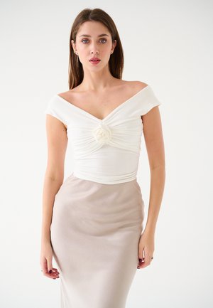Weißes Off-Shoulder-Top mit geknoteter Vorderseite und Stoffrose, kombiniert mit einem beigen, figurbetonten, glatten Rock. Einfaches und elegantes Design.