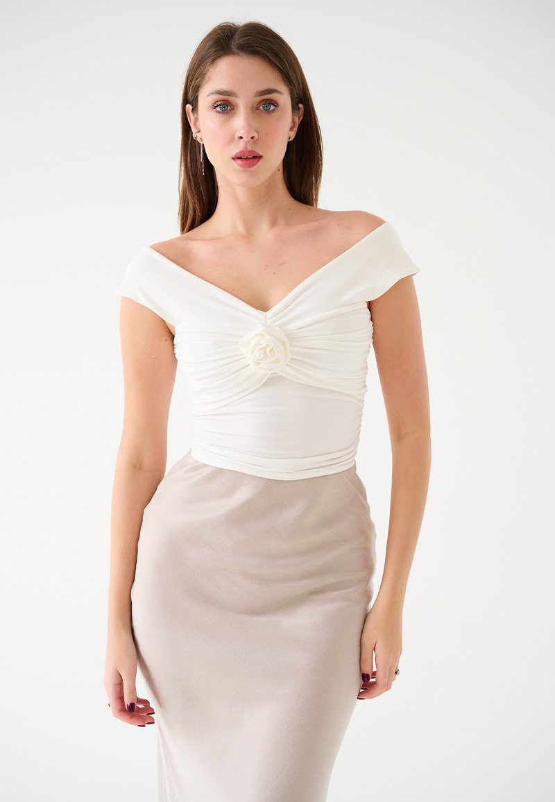 Top bianco scollato con un davanti annodato e dettaglio in rosa di stoffa, abbinato a una gonna aderente e liscia beige. Design semplice ed elegante.