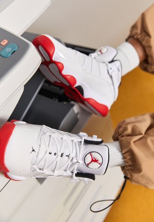 Baskets montantes blanches et rouges avec logos Jumpman portées avec un pantalon beige et des chaussettes blanches, assis près d'une imprimante.