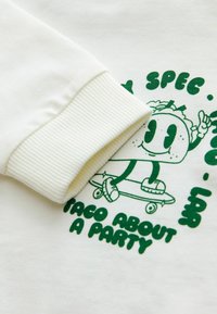 Vit bomullströja med grön grafisk design som föreställer en tecknad taco-karaktär på en skateboard, samt ribbade ärmslut.