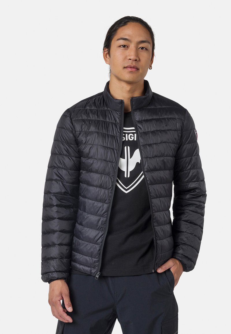 Svart puffjacka med kviltad design, hög krage och blixtlås framtill. Bärs över en svart t-shirt med ett grafiskt logotyp.