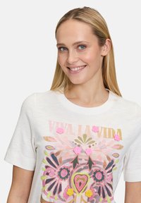 Weißes kurzärmliges T-Shirt mit lebhaftem Blumenmuster, das mit strukturierten rosa und grünen Akzenten sowie dem Text "Viva La Vida" versehen ist.