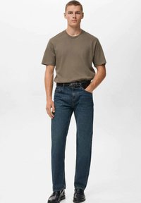 Ung mand iført en olivengrøn T-shirt, mørkeblå jeans med et sort bælte og sorte sko, stående med den ene hånd i lommen.