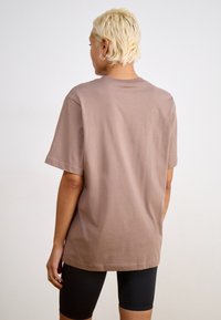 Rjav kratki rokav T-shirt iz mehkega bombaža z sproščenim krojem, viden zadaj, v kombinaciji s črnimi oprijetimi kratkimi hlačami.