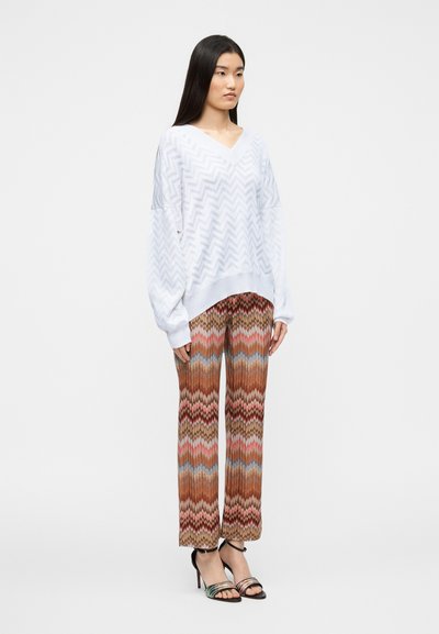 Λευκό, χαλαρά πλεκτό πουλόβερ με λαιμόκοψη σε σχήμα V και σχέδιο ζιγκ-ζαγκ· καφέ παντελόνι φαρδύ στο κάτω μέρος με ρίγες ζιγκ-ζαγκ σε κόκκινο, μπλε και μπεζ· μαύρα σανδάλια με τακούνι.