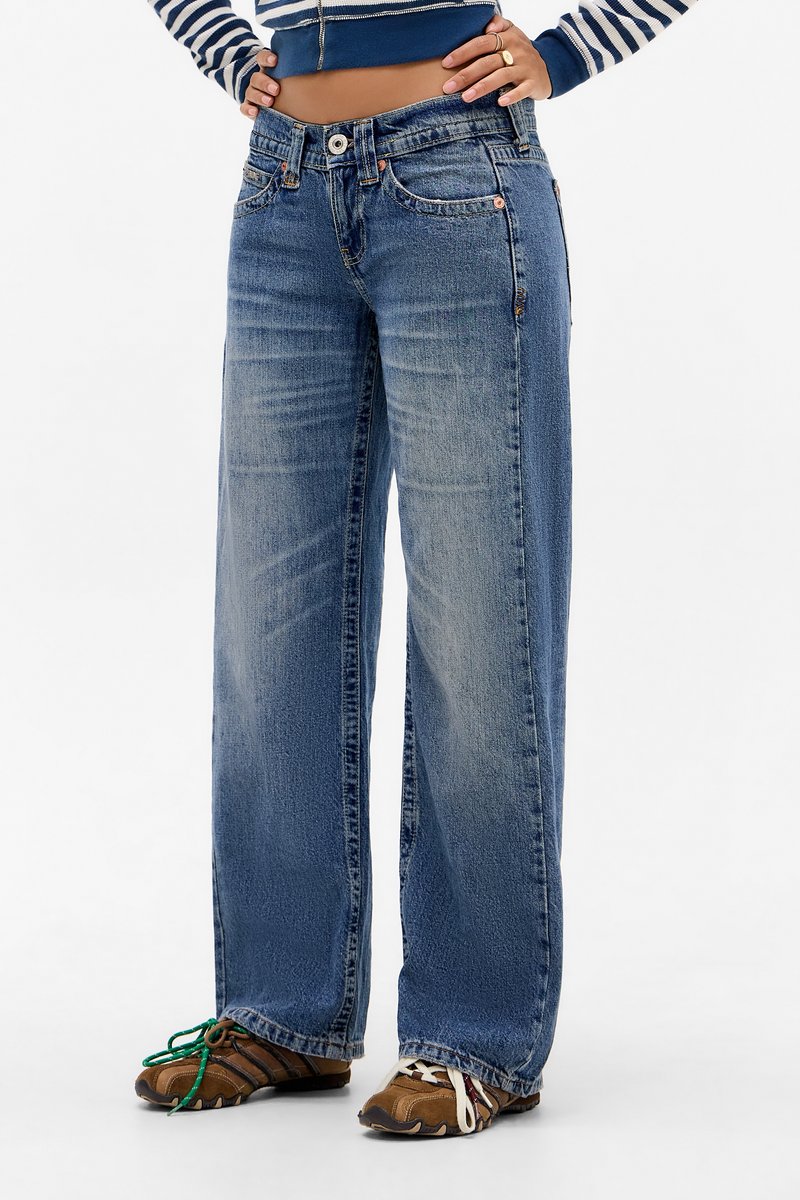 Person wearing loose blue jeans, brown athletic shoes with mismatched green and white laces, and a cropped striped top with hands on hips.