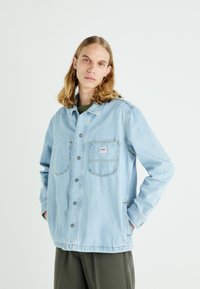 Veste en denim bleu clair avec des manches longues, fermeture à boutons, deux poches poitrine et une coupe décontractée. Texture douce, détails de couture visibles.