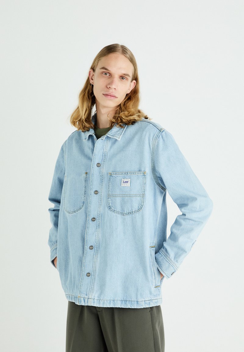 Veste en denim bleu clair avec des manches longues, fermeture à boutons, deux poches poitrine et une coupe décontractée. Texture douce, détails de couture visibles.