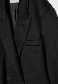 Gros plan d’un manteau noir en laine à double boutonnage avec un col cranté et trois boutons visibles sur le devant.
