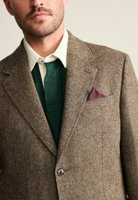 Brown tweed blazer with a notched lapel, brown button, and a chest pocket holding a maroon pocket square. Underneath, a green garment and white collar.