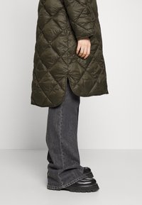 Olivově zelený prošívaný kabát s vysokým límcem, postranními rozparky a lesklou texturou, doplněný o šedé rozšířené džíny a objemné černé boty.