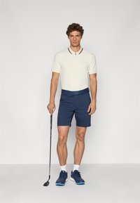 Polo majica u krem boji s tamnocrvenim ovratnikom, uparena s mornarskim kratkim hlačama. Model drži golf palicu i nosi mornarske tenisice.