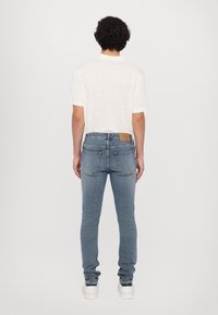 Muž stojící zády, oblečený v bílém tričku s krátkým rukávem, světle modrých slim-fit džínách a bílých teniskách, na bílém pozadí.