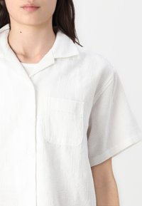 White short-sleeve shirt with a collared neckline, featuring a textured pattern, a chest pocket, and a button closure.
