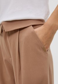 Pantalons beige ajustés avec une texture lisse, dotés de poches latérales et d'un petit accent doré sur la poche.
