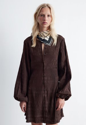 Brown button-down dress with textured fabric, long puff sleeves, and a loose fit. A black and white scarf is worn around the neck.