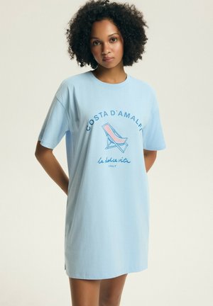 Kobieta o kręconych włosach, ubrana w jasnoniebieską, oversize'ową sukienkę typu T-shirt z grafiką "Costa d'Amalfi" i leżakiem na przodzie.