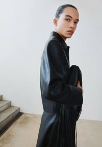 Czarna skórzana kurtka o wyraźnej strukturze, z wysokim kołnierzem i ozdobą z frędzli. Gładka faktura, dopasowana w talii.