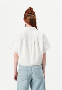 Persona con i capelli rossi intrecciati e raccolti in un bun, che indossa una camicia bianca a maniche corte larga e jeans azzurri a vita alta, fotografata da dietro.