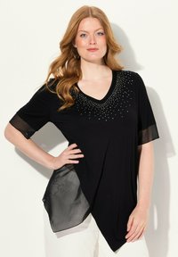 Haut noir avec un col en V et de courtes manches en maille transparente. Présente des embellissements en perles le long de l'encolure et un ourlet asymétrique.